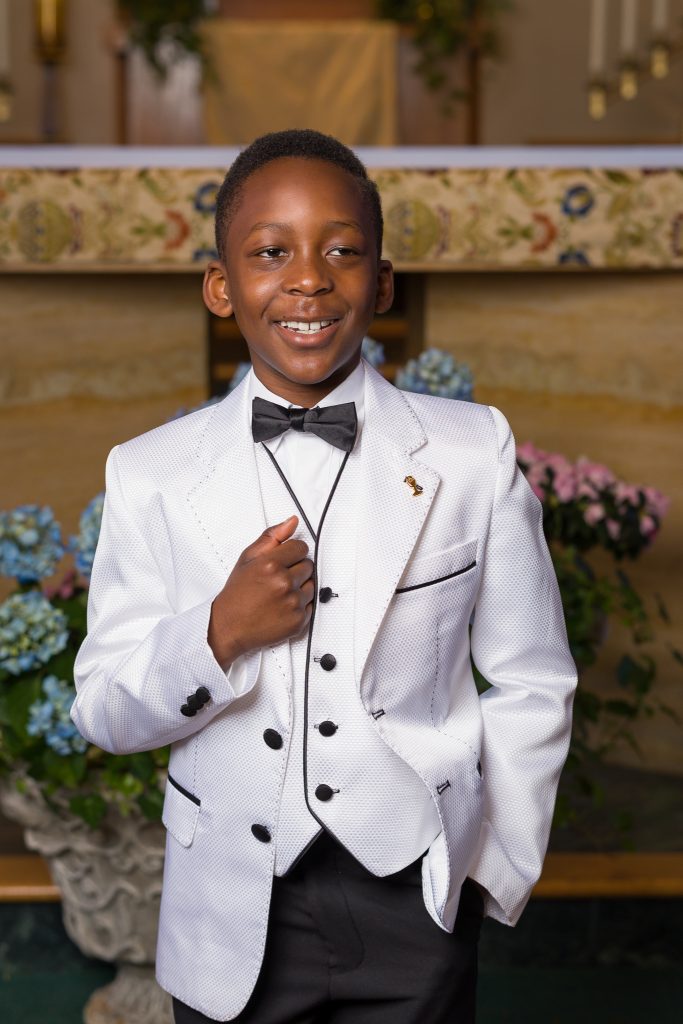 Boy in a white tuxedo jacket and bow tie smiling in a church, First Communion portrait in St. Bernadette's Church, Northborough, MA