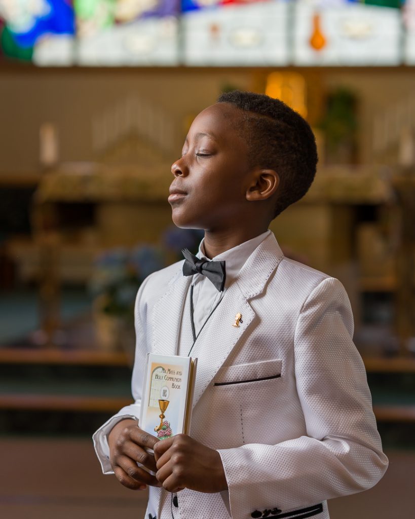 An African American little boy inside a church holding a First Communion book with his head tilted up towards the light and his eyes closed, First Communion portrait session in Northborough, MA.
