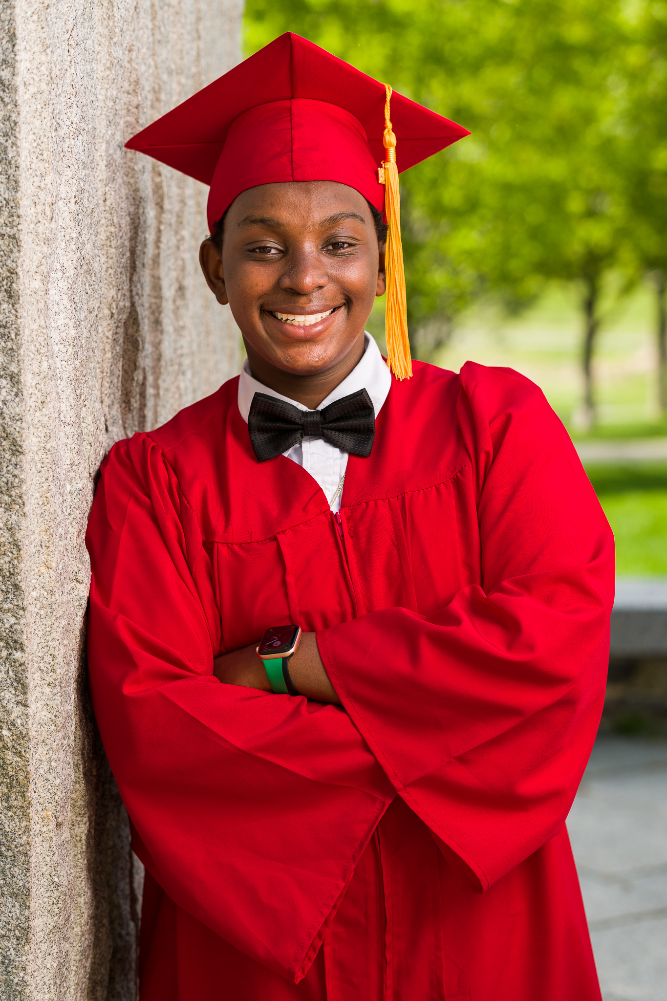 Graduate in a red cap and gown leaning against a stone wall, graduation portrait in Greenhill Park, Worcester, MA