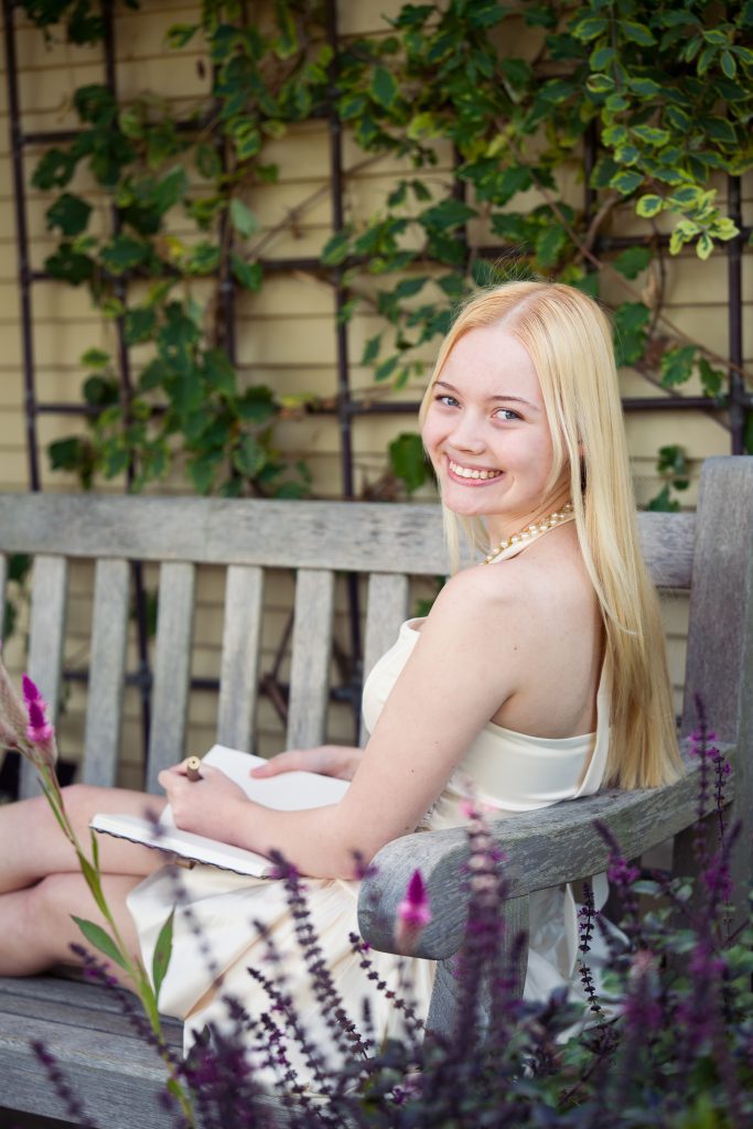 High school girl sitting on a bench in a garden drawing in a book and looking over her shoulder at the camera, senior portrait session in Boylston, MA.