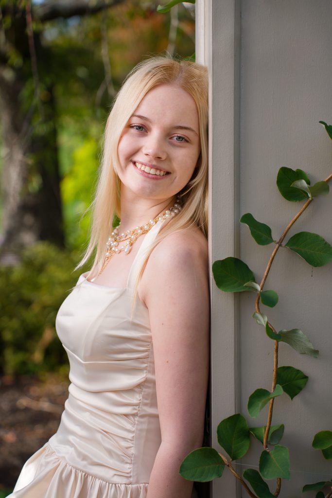 Smiling senior girl in a champagne dress leaning against a vine-covered column, senior portrait in Boylston, MA