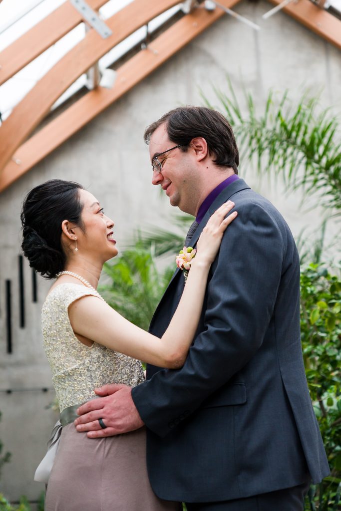 A wedding photo of a Southeast Asian woman and Caucasian man looking at each other in an embrace inside a conservatory at the New England Botanic Garden. A wedding portrait session in Boylston, MA.