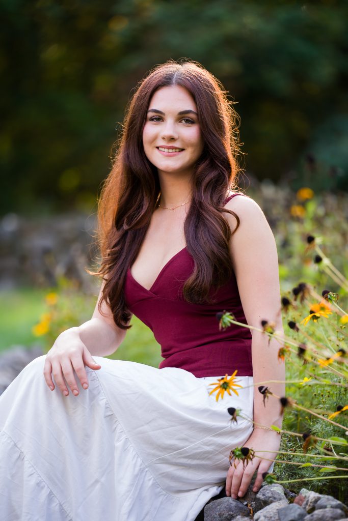 Senior girl in a burgundy top and white skirt sitting among wildflowers, senior portrait in Garden of Sweet Remembrance, Shrewsbury, MA