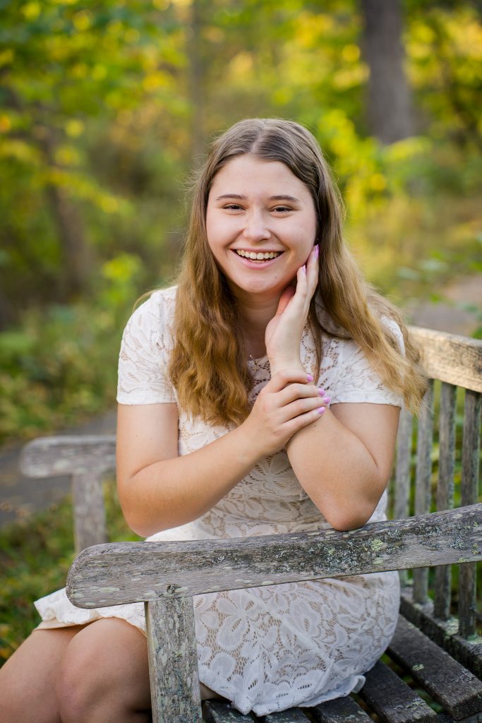 Smiling senior girl in a white lace dress sitting on a wooden bench, senior portrait in Acton Arboretum, Acton, MA