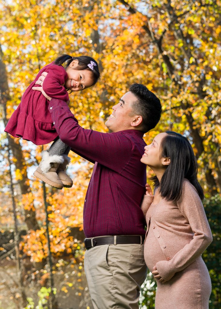 A Southeast Asian family of three where the little girl is held up in the air by the father and her mother is looking up at her behind the father, family portrait session in Westborough, MA.