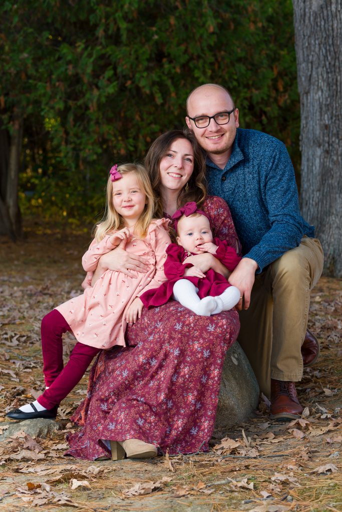 A family of four in a wooded setting where the mother is sitting on a rock holding a baby and a toddler is leaning against her, and the husband kneeling on one knee in a close embrace, family portrait session in Shrewsbury, MA.