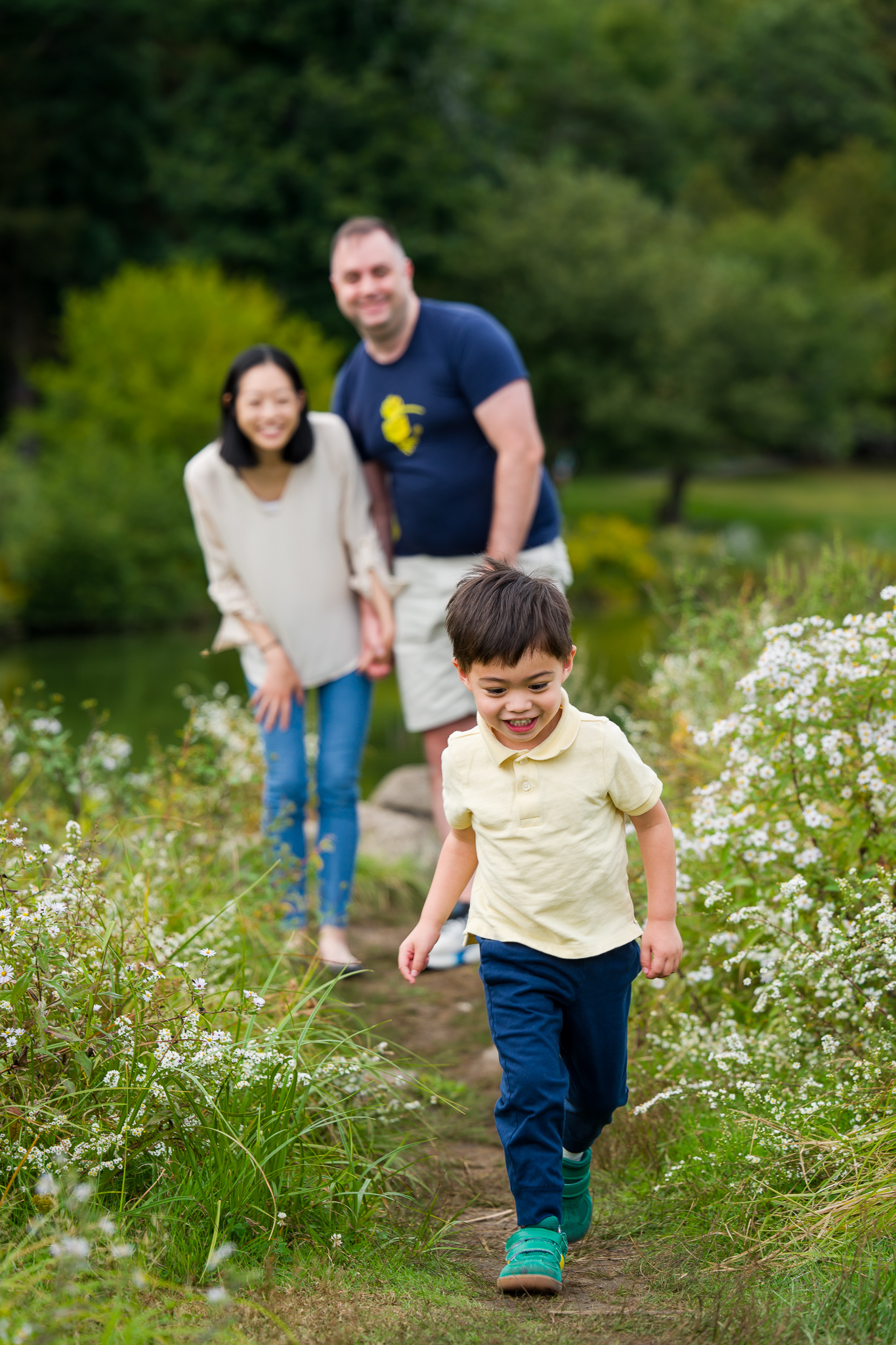 A family of three where a little boy is running towards the camera while his parents are blurred in the background watching him and smiling.