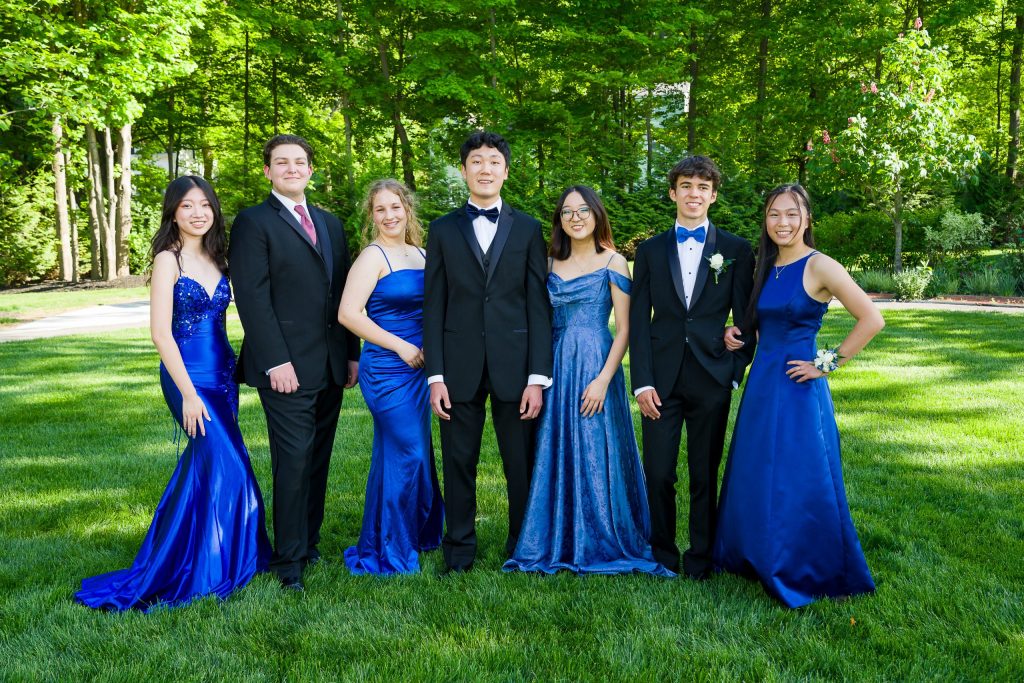 Seven teens in formal blue gowns and tuxedos standing on a lawn, prom group portrait in Northborough Town Common, Northborough, MA
