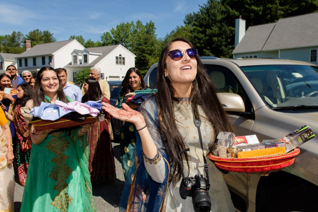 Woman with a camera cheering as guests in saris carry gift baskets outdoors, Indian celebration in Shrewsbury, MA