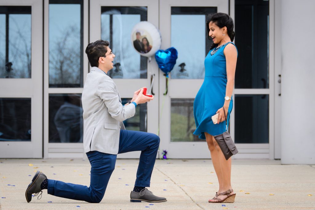 Man proposing on one knee to a woman in a blue dress outside glass doors, proposal photography in Shrewsbury High School, Shrewsbury, MA