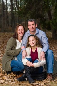 Parents and daughter sitting close together in autumn leaves, family portrait session in Dean Park, Shrewsbury, MA