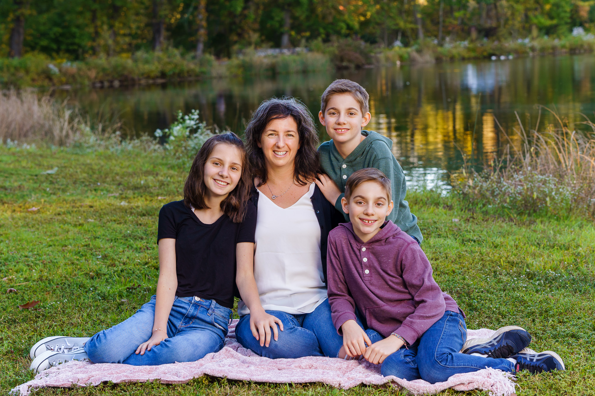 Mother and three children sitting on a picnic blanket by a pond, family portrait session in Dean Park, Shrewsbury, MA