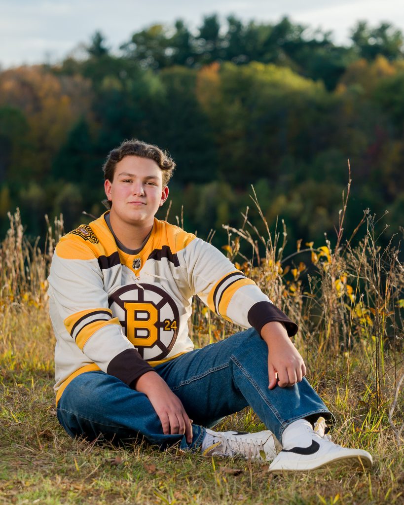 Senior guy in a hockey jersey sitting in a field of tall grass, senior portrait in Old Stone Church, West Boylston, MA