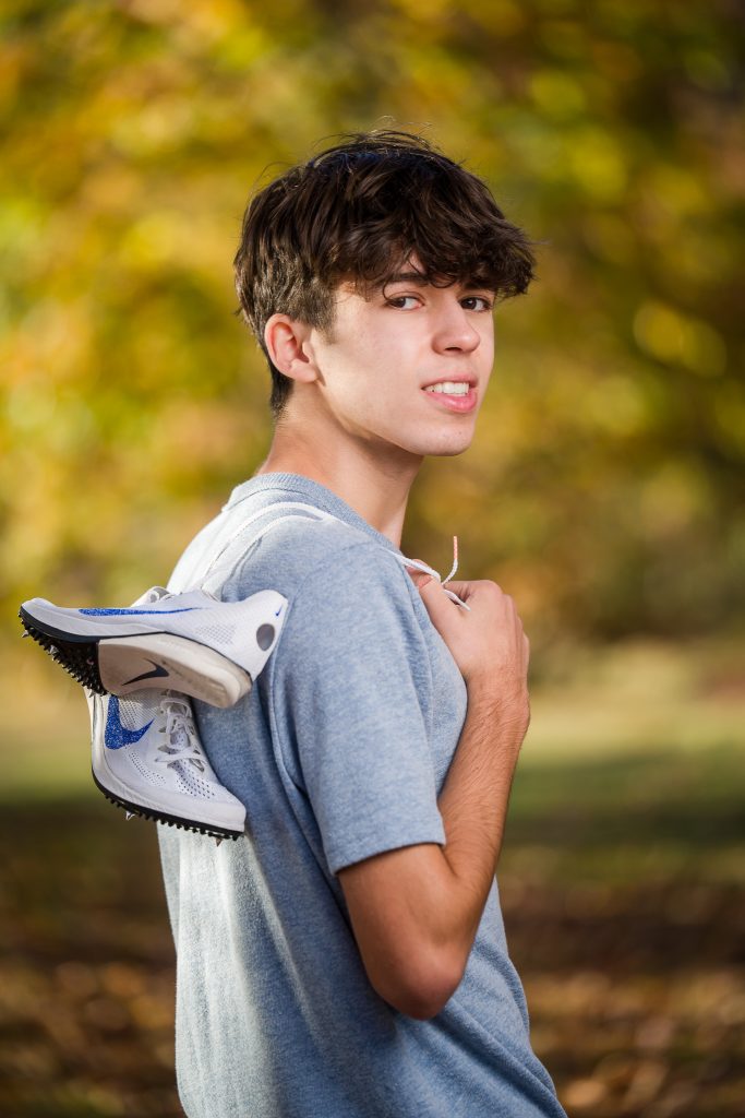 Senior guy in a gray tee with track shoes slung over his shoulder, autumn senior portrait in West Boylston, MA