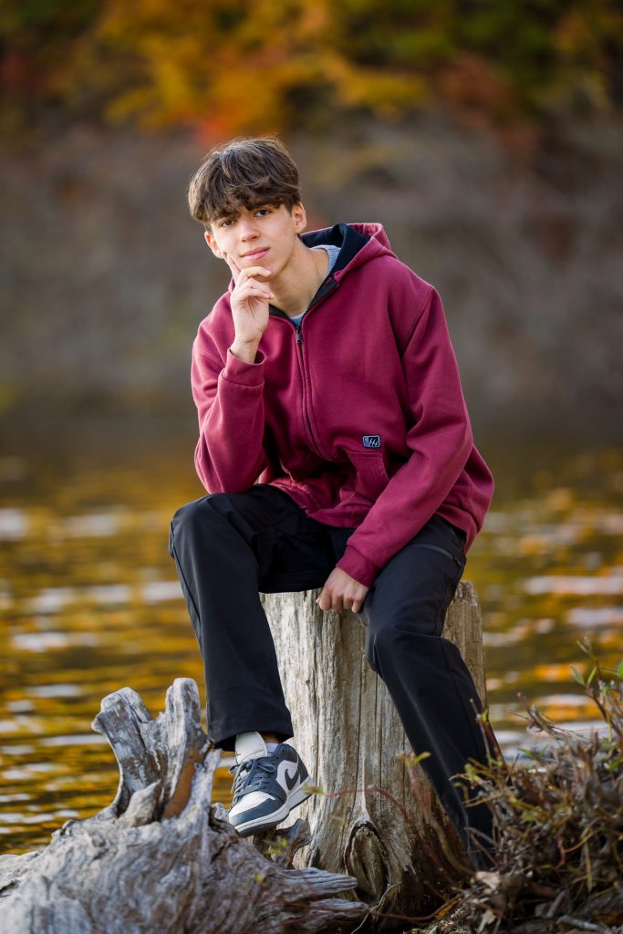 Senior guy in a maroon hoodie sitting on a tree stump by the water, senior portrait in Old Stone Church, West Boylston, MA