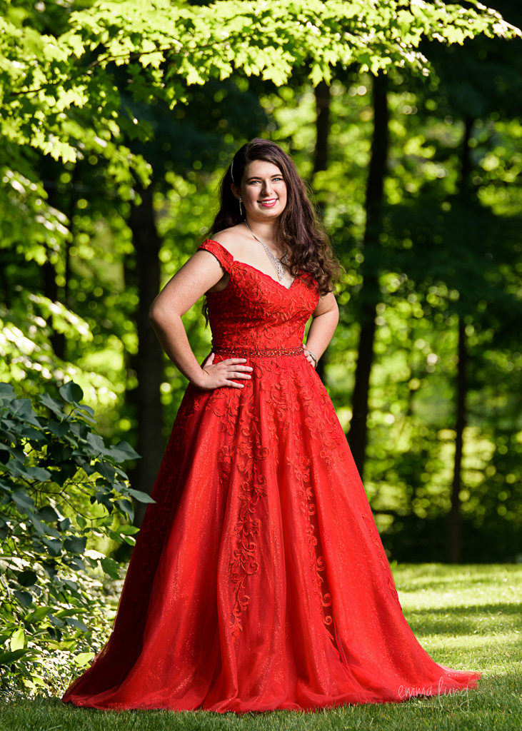 A high school girl in a red prom dress posing with tree leaves backlit behind her, a prom portrait session in Shrewsbury, MA.