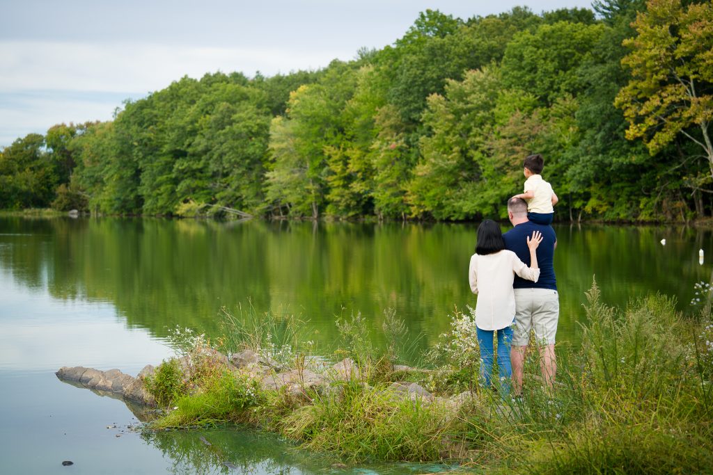 A family of three where the toddler is sitting on the dad's shoulder and the mother is close beside them all looking out at a body of water, family portrait session in Shrewsbury, MA.
