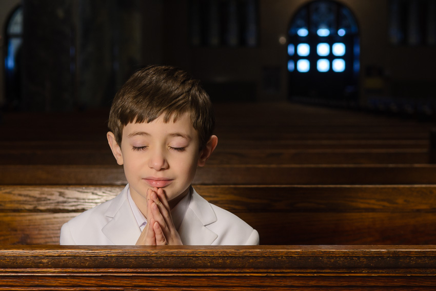 Boy in a white jacket praying with eyes closed in a church pew, First Communion portrait in St. Catherine of Genoa Church, Somerville, MA