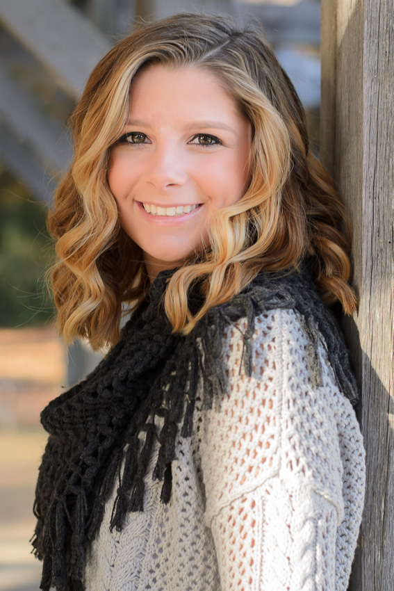 Smiling senior girl with wavy hair and a black scarf leaning on a wooden post, senior portrait in Moore State Park, Paxton, MA