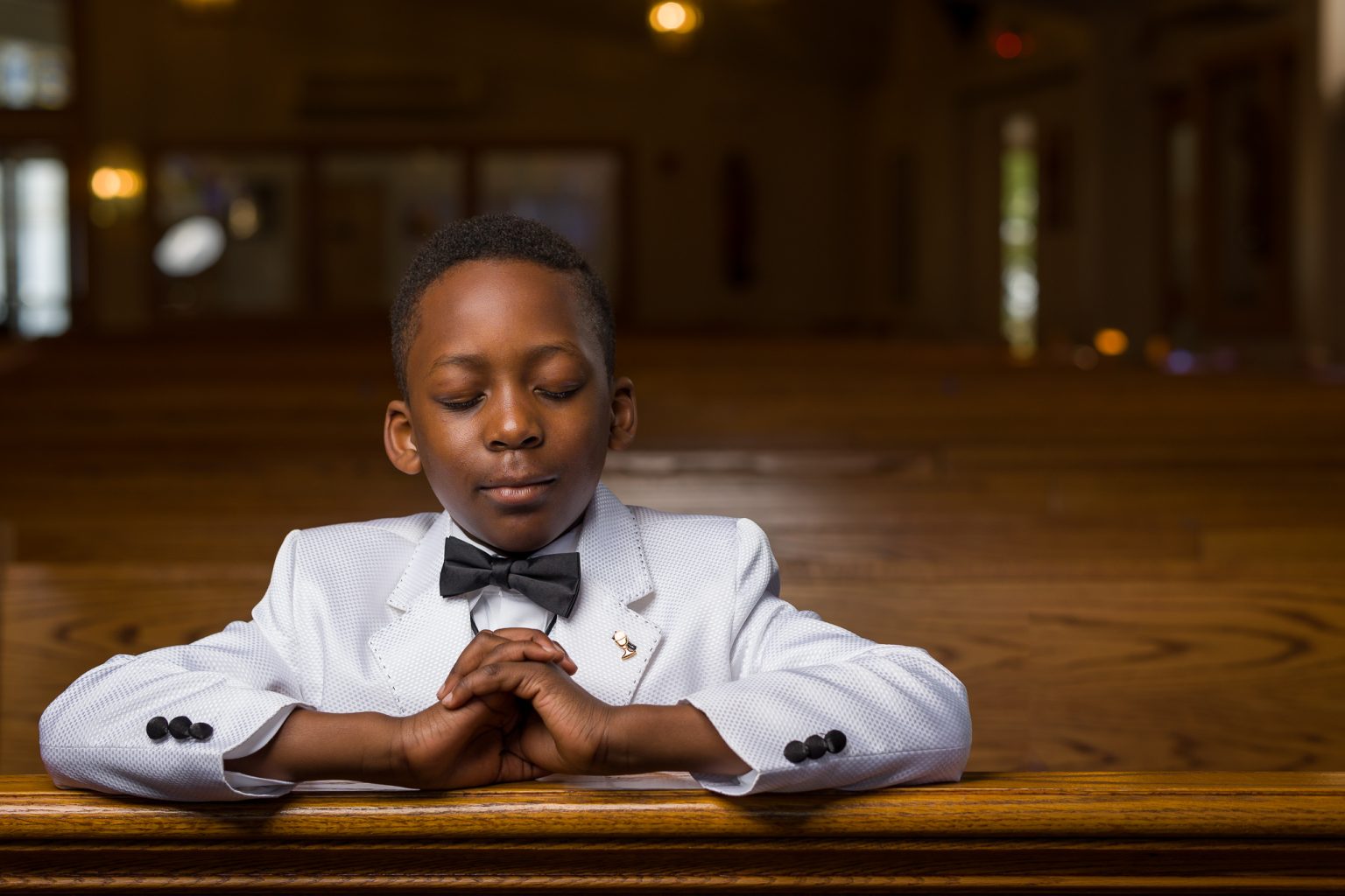 Boy in a white suit and bow tie praying in a church pew, First Communion portrait at St. Bernadette's, Northborough, MA