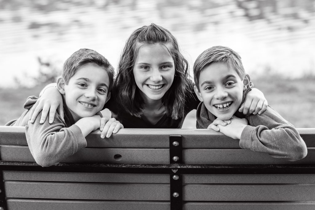 Black-and-white portrait of three siblings smiling over a park bench, family portrait in Dean Park, Shrewsbury, MA