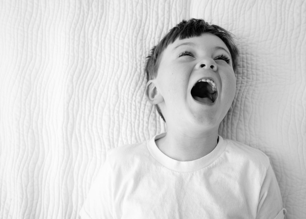 Black-and-white portrait of a laughing boy lying on white bedding, family portrait session at a client's home in Northborough, MA