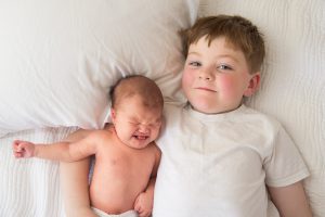 Newborn baby lying beside smiling big brother on white bedding, family portrait session at a client's home in Northborough, MA