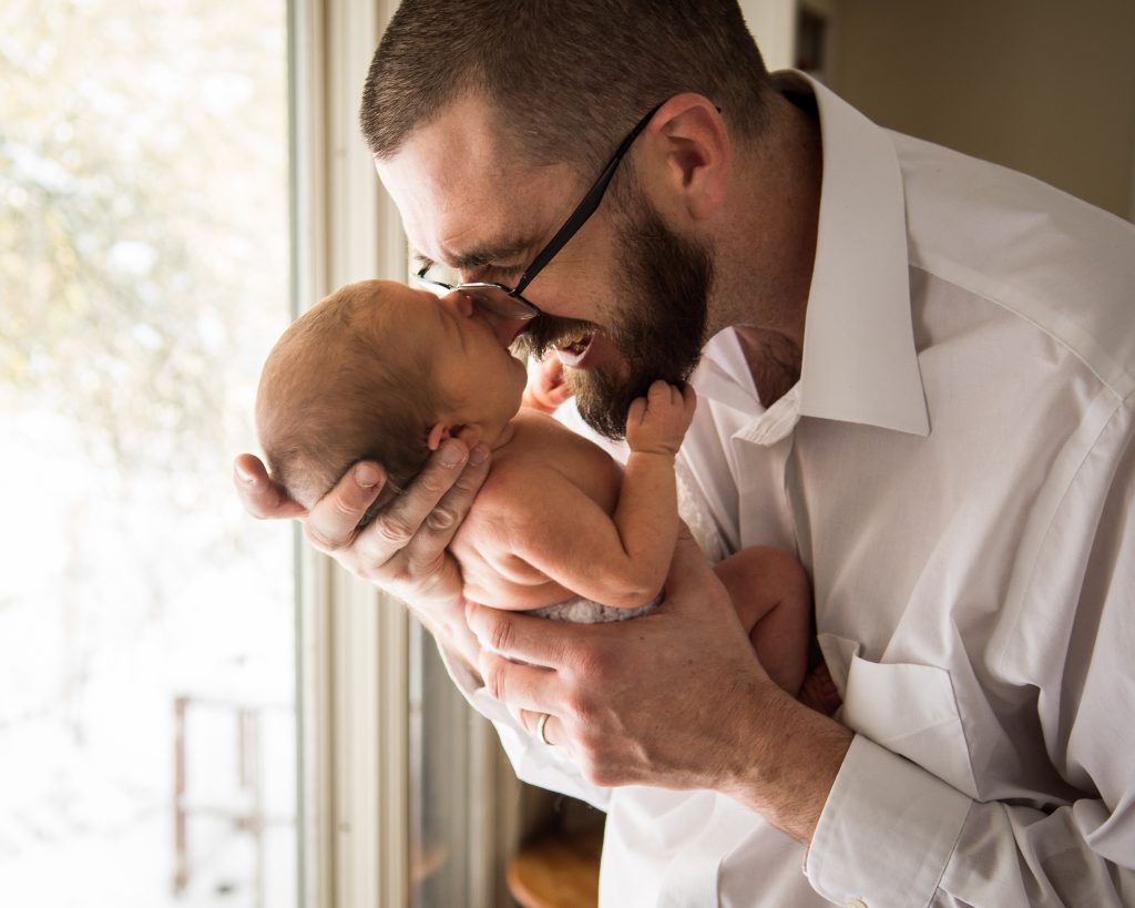 Father in glasses kissing his newborn baby by a window, newborn family portrait at a client's home in Northborough, MA