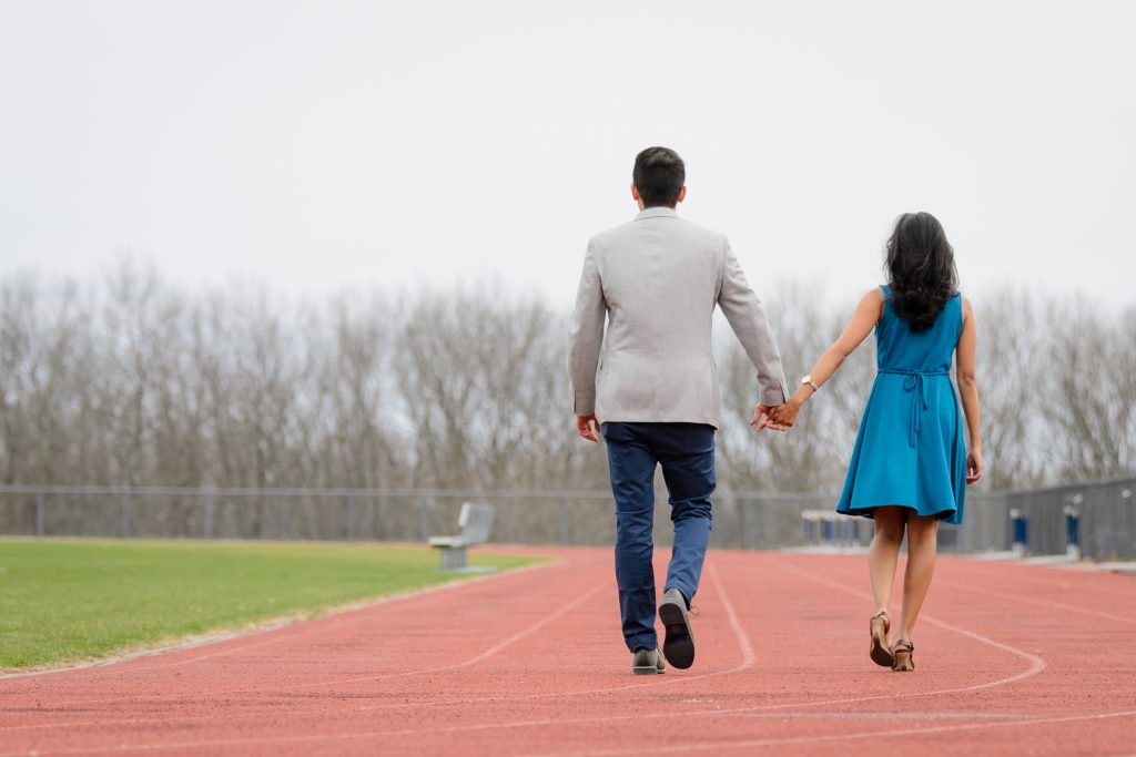 A South Asian man and woman holding hands and walking away from the camera on a running track where the couple met years ago, engagement portrait session in Shrewsbury, MA. 